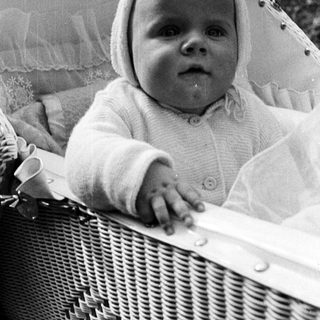 Here's a brief summary of the image: Baby in a basket, wearing a bonnet and sweater.