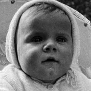 Here's a brief summary of the image: Baby in a basket, wearing a bonnet and sweater.