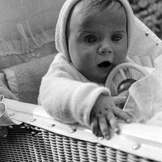 Baby in a basket, wearing a bonnet, looking forward.
