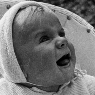 Baby in a hooded sweater sits in a wicker chair.