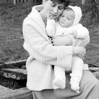 Here's a brief summary of the image: Woman holds baby outdoors, sitting on a wooden structure.