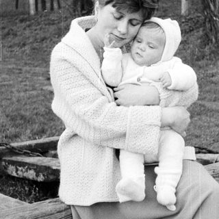 Here's a brief summary of the image: Woman holds baby outdoors, sitting on a wooden structure.
