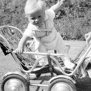 Here's a brief summary of the image: Baby standing in vintage stroller outdoors, looking away.