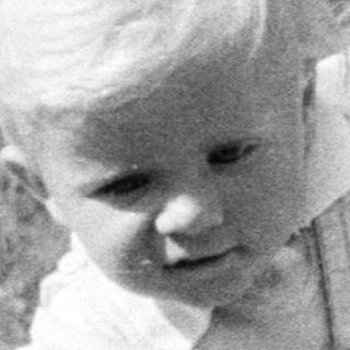 Here's a brief summary of the image: Baby standing in vintage stroller outdoors, looking away.