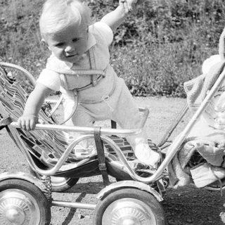 Here's a brief summary of the image: Baby standing in vintage stroller outdoors, looking away.