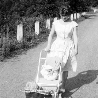 Here's a brief summary of the image: Young woman walks along a road, pushing a baby carriage.