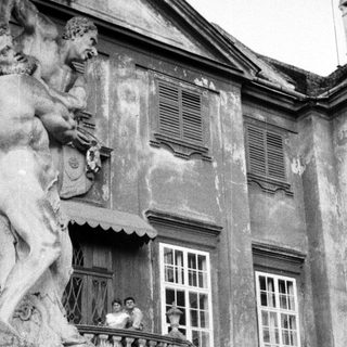 Here's a brief summary of the image: People on a balcony overlooking a grand, aged building.
