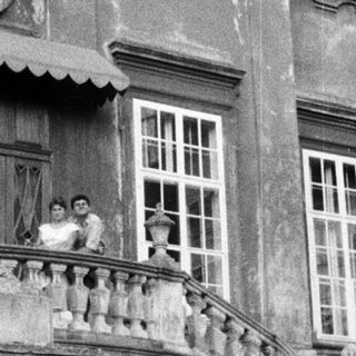 Here's a brief summary of the image: People on a balcony overlooking a grand, aged building.