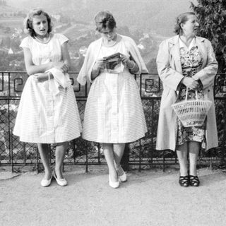 Three women in vintage dresses stand near a fence.