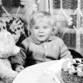 Child with teddy bear sits near birthday cake.