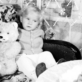 Baby sits with teddy bear in vintage chair.