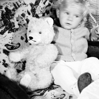 Baby sits with teddy bear in vintage chair.