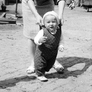 Here's a brief summary of the image: Woman supports a toddler taking first steps outdoors.