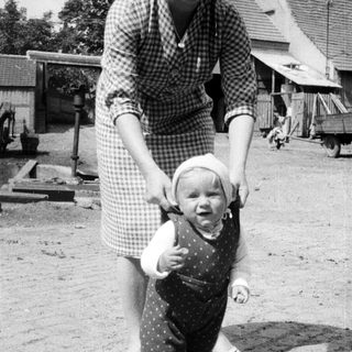Here's a brief summary of the image: Woman supports a toddler taking first steps outdoors.