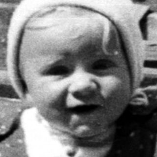 Baby in overalls walks with an adult outdoors.