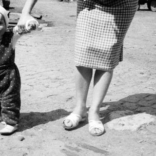Baby in overalls walks with an adult outdoors.