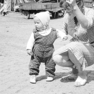 Baby takes first steps near a farmhouse, woman watching.