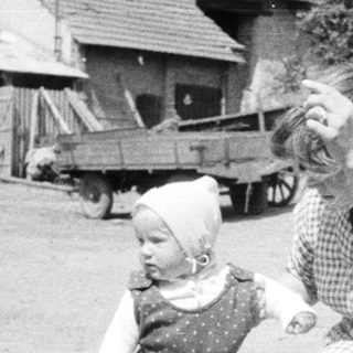 Baby takes first steps near a farmhouse, woman watching.