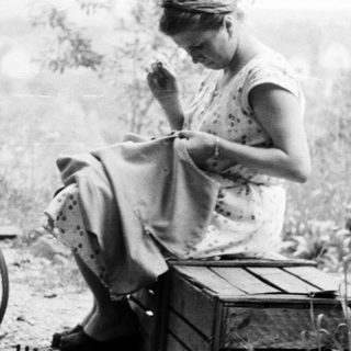 Woman sits outdoors, sewing in a garden setting.