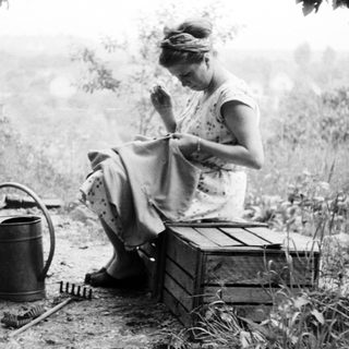 Woman sits outdoors, sewing in a garden setting.