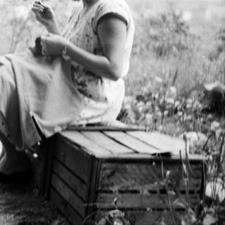 Here's a brief summary of the image: Woman sits on crate, sewing in a garden setting.
