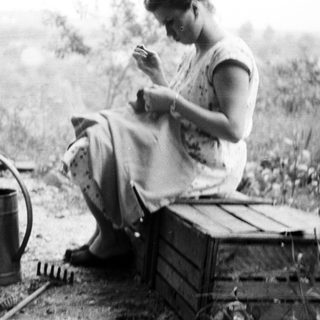 Here's a brief summary of the image: Woman sits on crate, sewing in a garden setting.