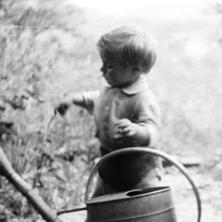 Child stands near watering can, woman sits on crate.