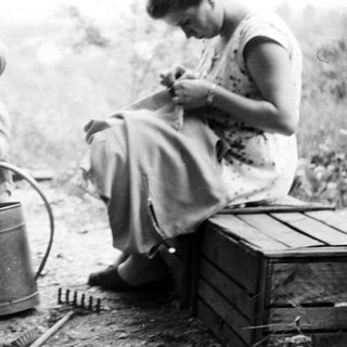 Child stands near watering can, woman sits on crate.