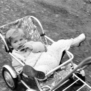 Here's a brief summary of the image: Baby sits in a vintage stroller outdoors.
