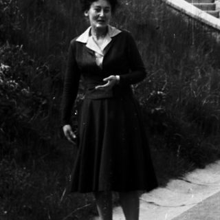 Woman stands on road with stairs and greenery behind.