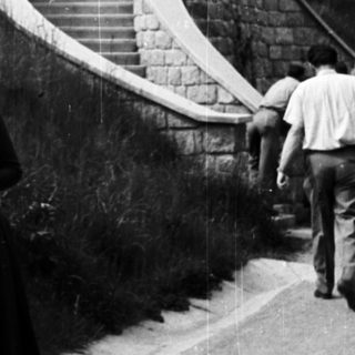 Woman stands on road with stairs and greenery behind.
