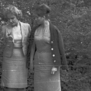 Two women in vintage dresses stand near foliage.