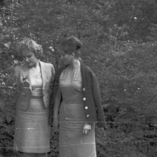 Two women in vintage dresses stand near foliage.