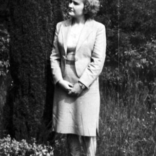 Woman stands in a garden with tall trees.