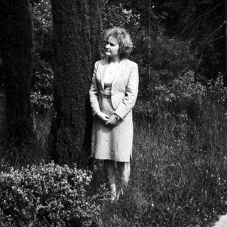 Woman stands in a garden with tall trees.
