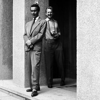 Here's a brief summary of the image: Two men in suits stand outside a building.
