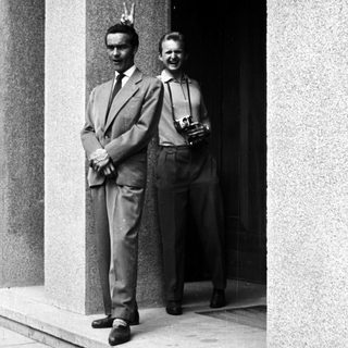 Here's a brief summary of the image: Two men in suits stand outside a building.