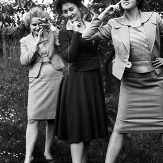 Here's a brief summary of the image: Three women in suits pose outdoors with playful gestures.
