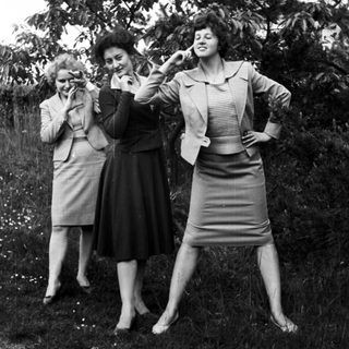Here's a brief summary of the image: Three women in suits pose outdoors with playful gestures.