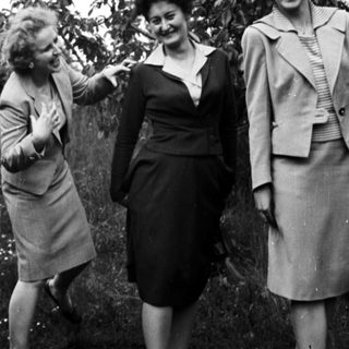 Three women in suits pose outdoors in a garden setting.