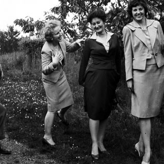 Three women in suits pose outdoors in a garden setting.