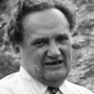Man in suit gestures in a hillside landscape.