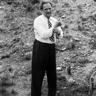 Man in suit gestures in a hillside landscape.