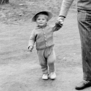 Man and child walk hand-in-hand outdoors in a wooded area.