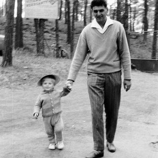 Man and child walk hand-in-hand outdoors in a wooded area.