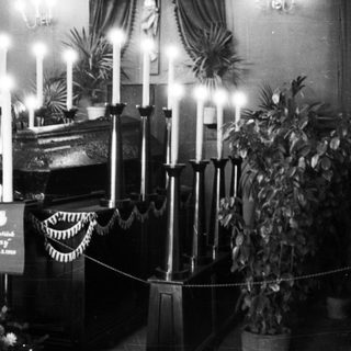 Here's a brief summary of the image: Funeral scene with a casket, candles, and foliage.