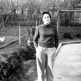 Woman stands in a yard with a chicken nearby.