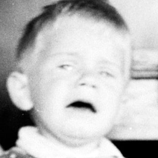 Infant in polka-dot overalls stands on a patterned rug.