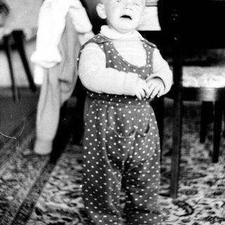 Infant in polka-dot overalls stands on a patterned rug.