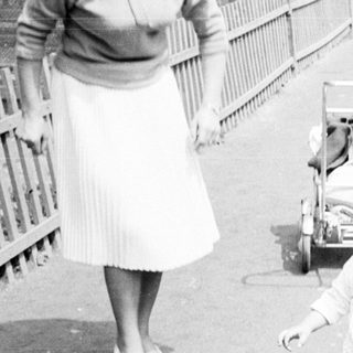 Baby in a suit walks near a woman and bench.